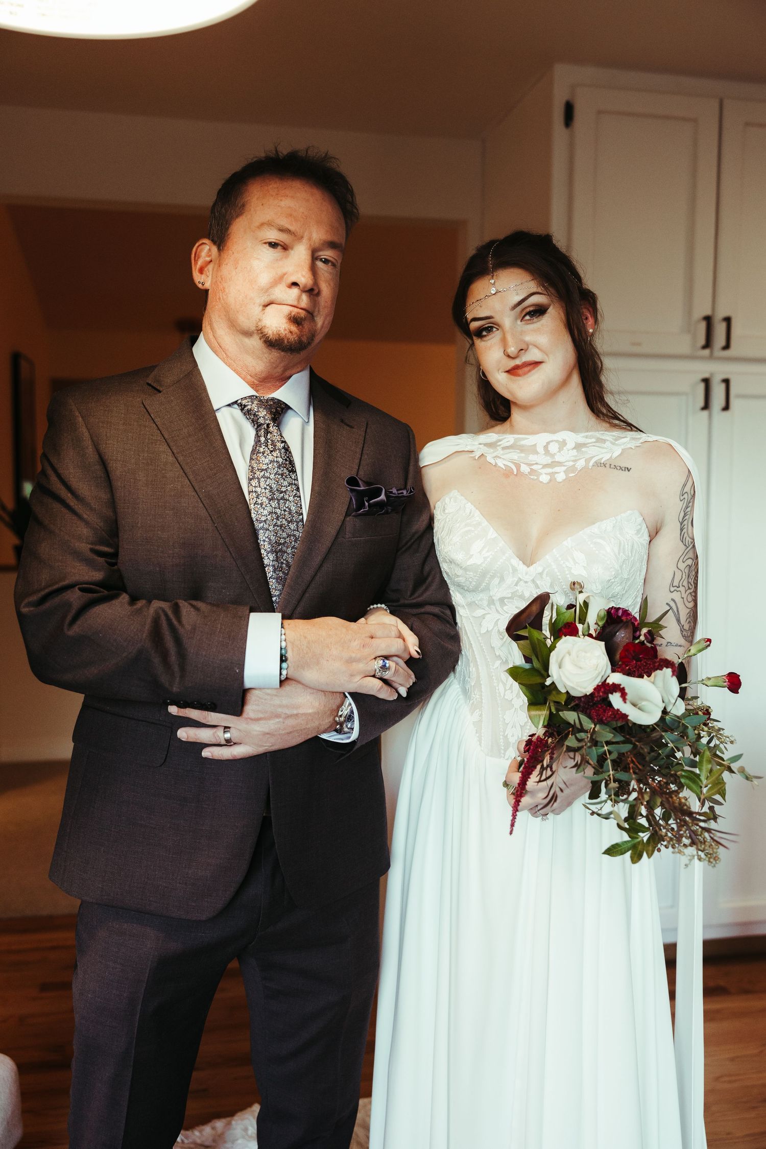 Bride in a lace wedding gown holding a burgundy and white bouquet stands beside a man in a brown suit and patterned tie.