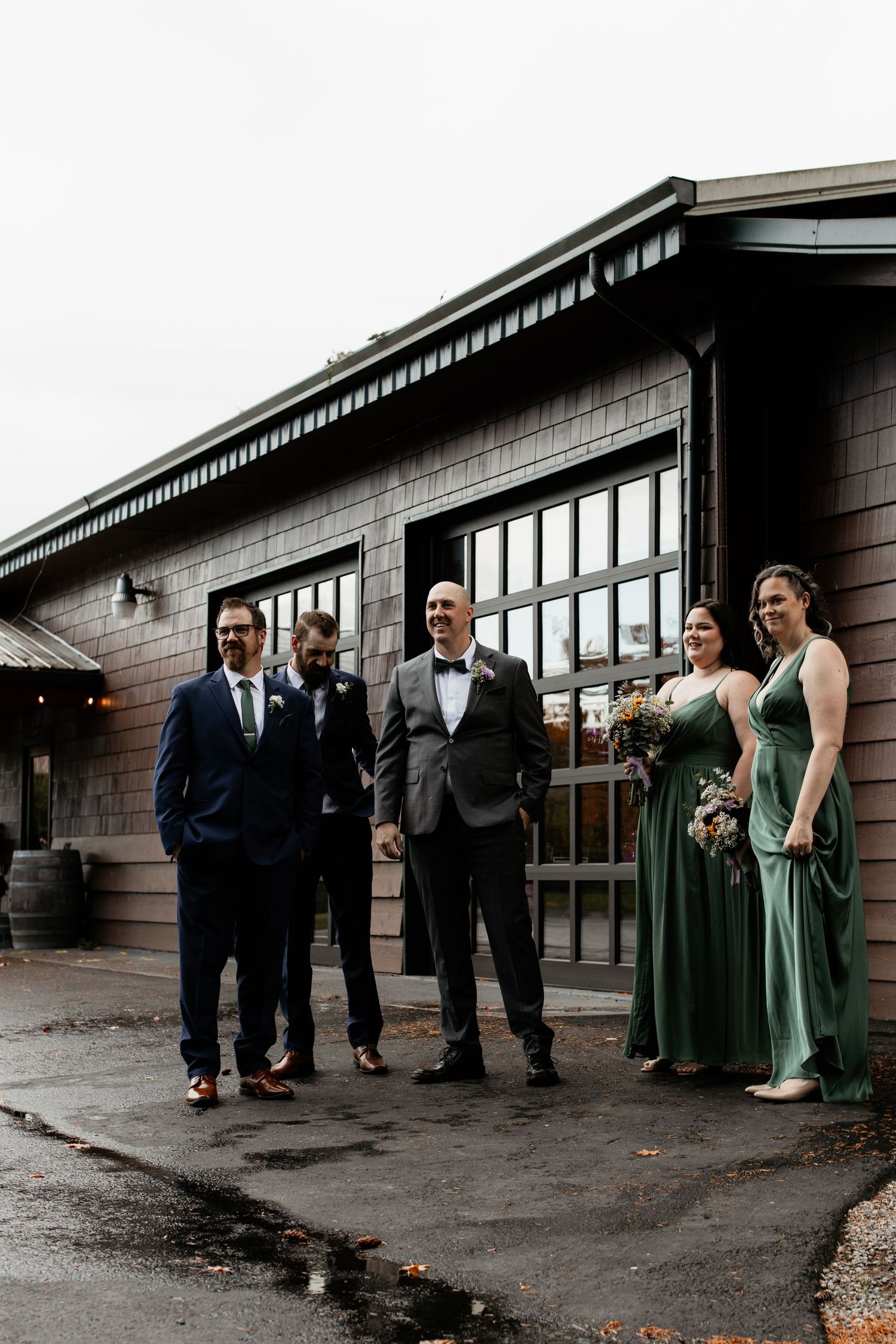 Wedding party poses outside rustic barn venue in sage green bridesmaid dresses and navy groomsmen suits.