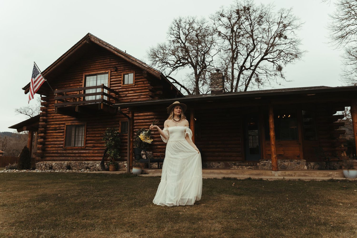 Bride in white gown stands before rustic log cabin with American flag, holding bouquet on overcast wedding day.