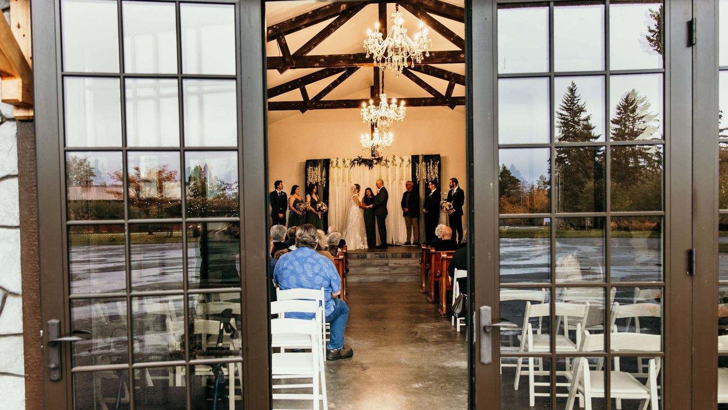 Wedding ceremony viewed through glass French doors in an elegant venue with chandeliers and white chairs.