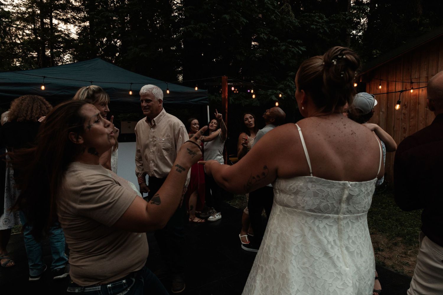 Two women dancing together at an outdoor evening wedding reception with string lights and guests in the background.