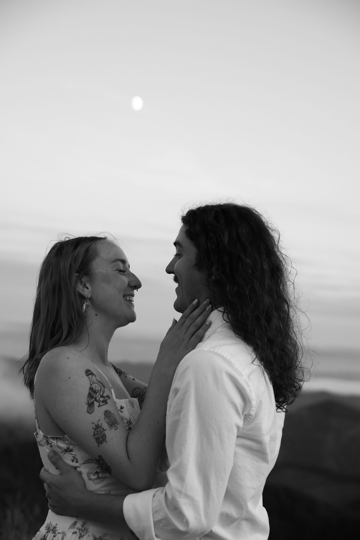 Black and white photo of a smiling couple embracing romantically under a full moon at dusk by the ocean.