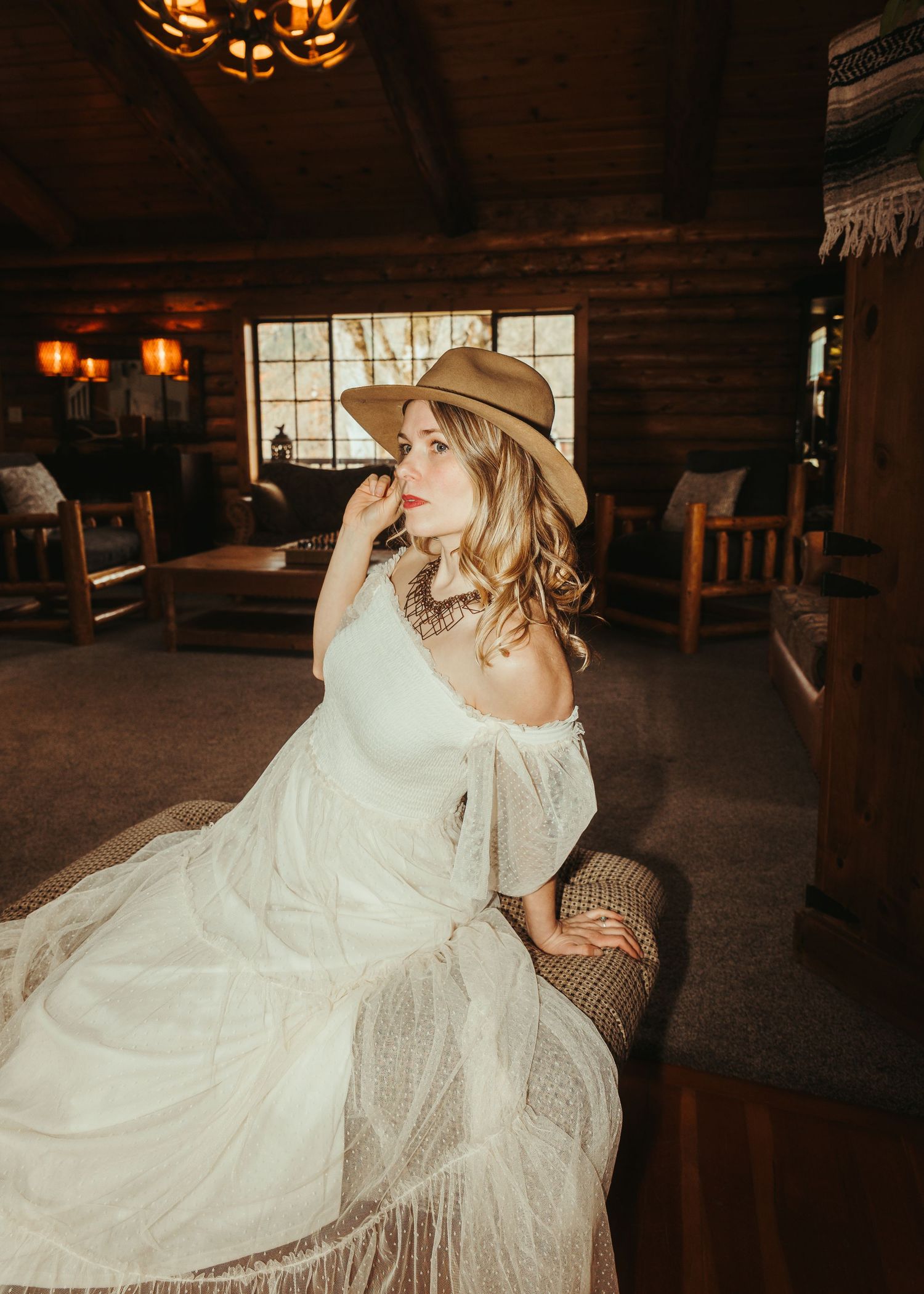 A bride in a white off-shoulder dress and tan cowboy hat sits elegantly in a rustic log cabin interior.