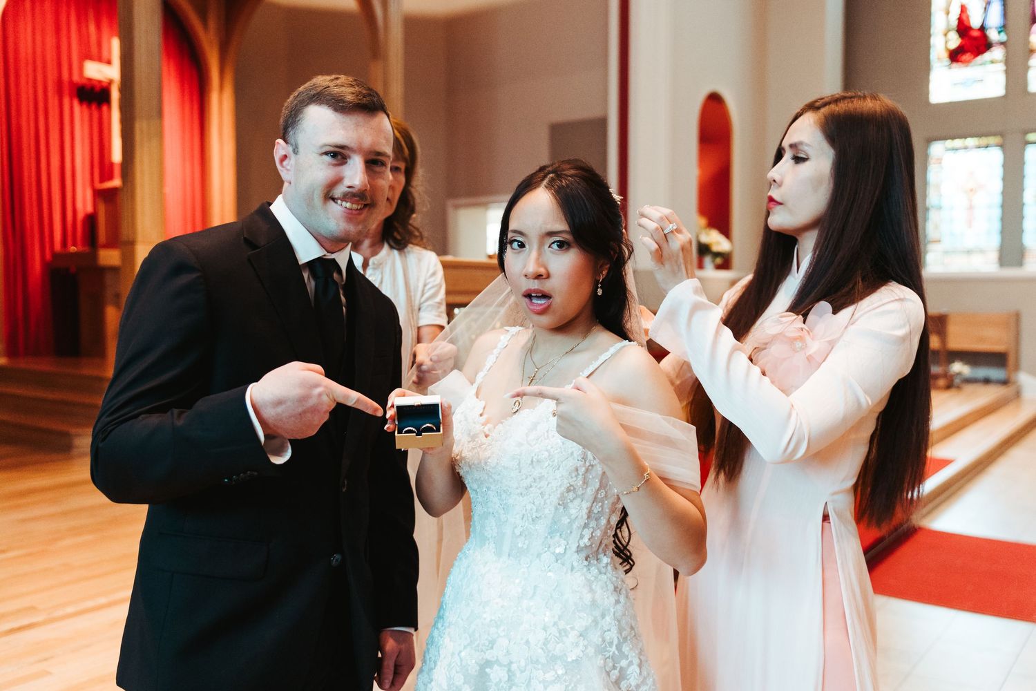 Groom in black suit and bride in white dress pointing at ring box with woman in white dress at church.