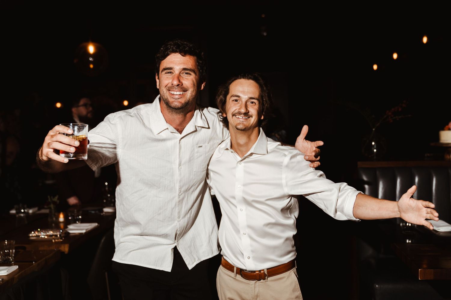 Two smiling men in white shirts celebrate at a dimly lit venue, one holding a champagne glass.