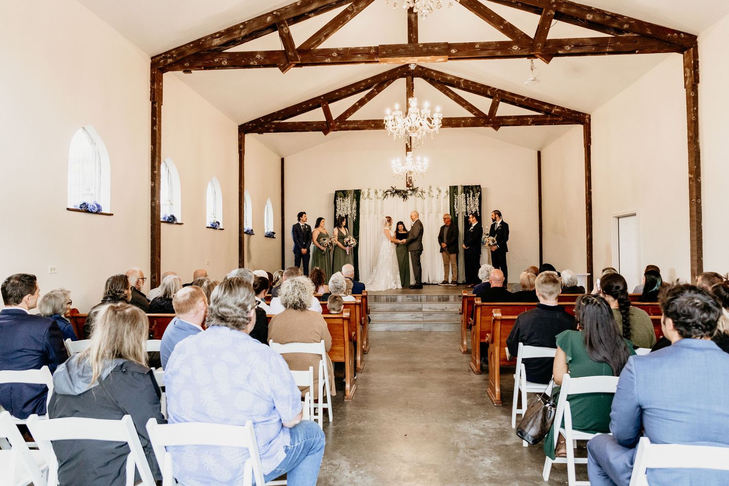 Guests seated in white chairs watch a wedding ceremony in a rustic chapel with wood beam ceilings.
