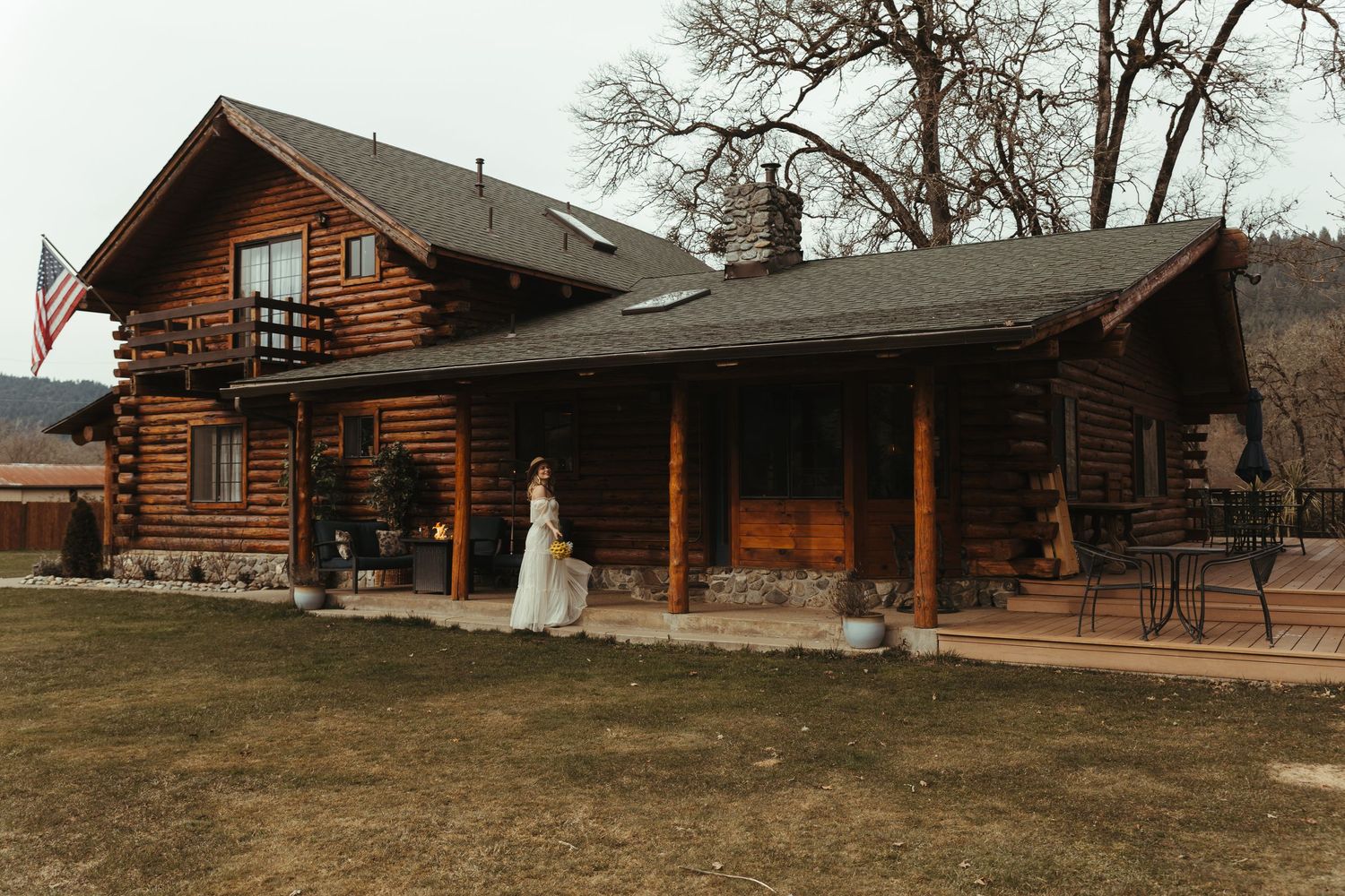 Bride in white wedding gown standing outside a rustic log cabin with an American flag.