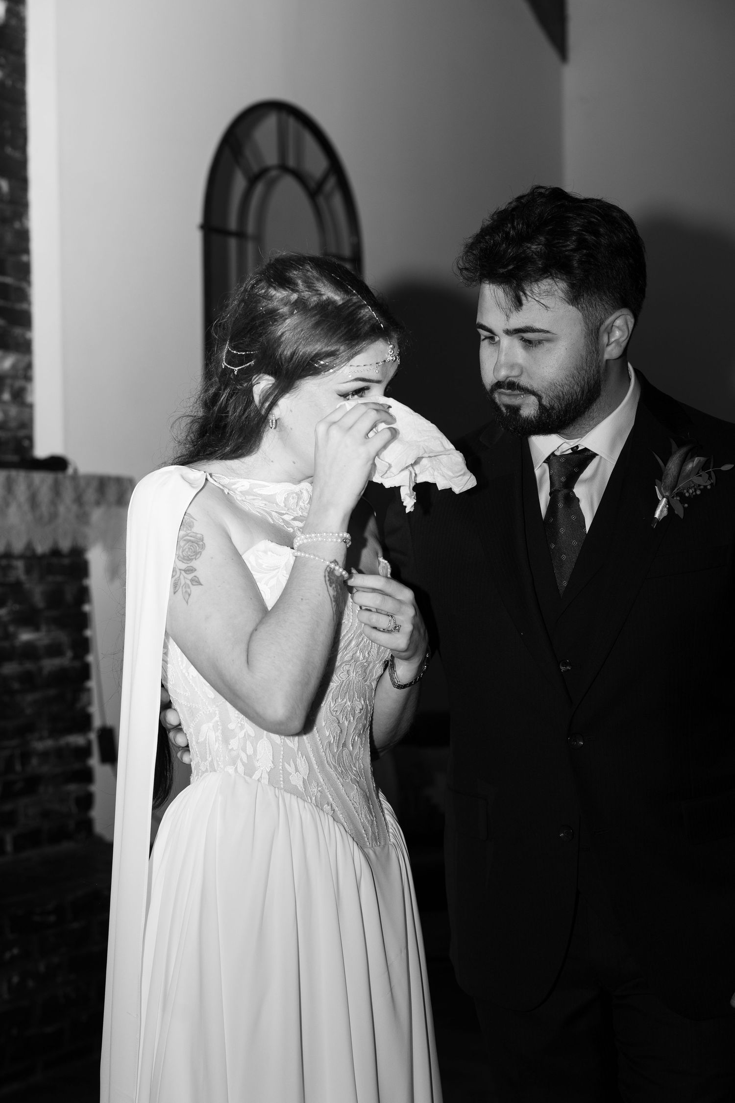 Black and white photo of an emotional bride dabbing her tears with a tissue while a bearded groom in a dark suit looks on at their wedding.