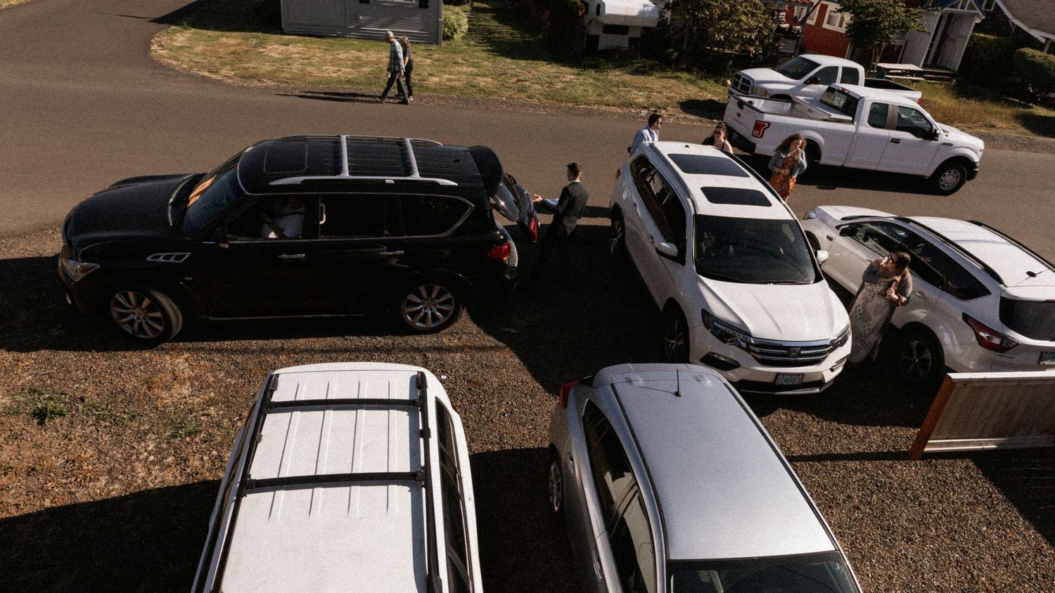 Aerial view of multiple cars parked in a driveway, including black SUV and white vehicles, with people standing nearby.