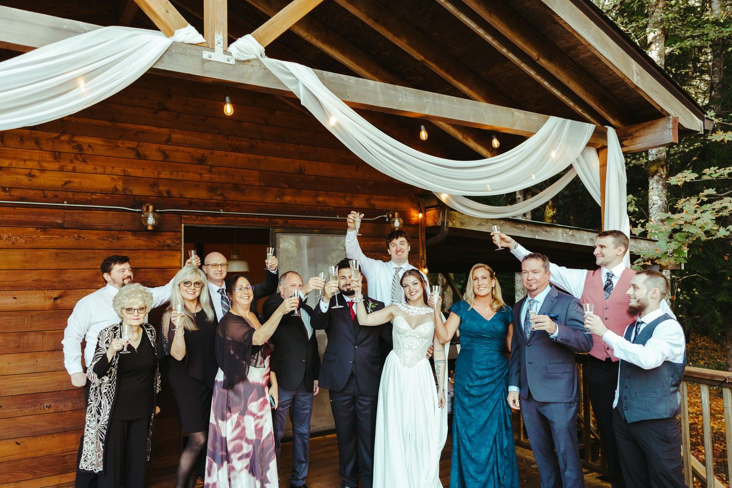 Bride and groom celebrate with cheering wedding guests under a rustic wooden pavilion draped in white fabric and string lights.