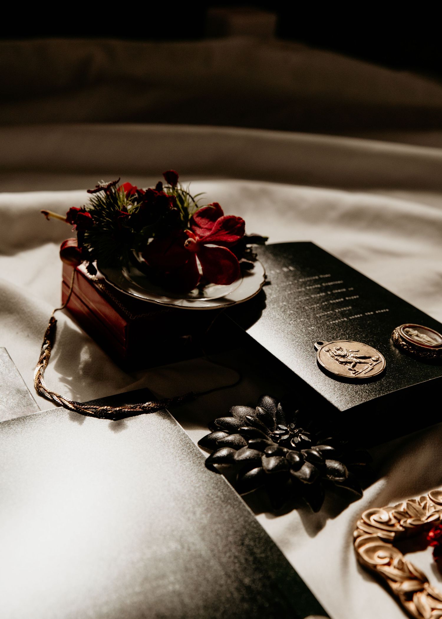 Dark elegant flat lay with red flowers, black book, gold medals, and decorative items on white fabric.