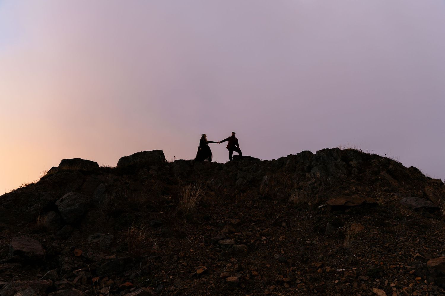 Silhouette of a couple holding hands on a rocky mountain summit at dusk.
