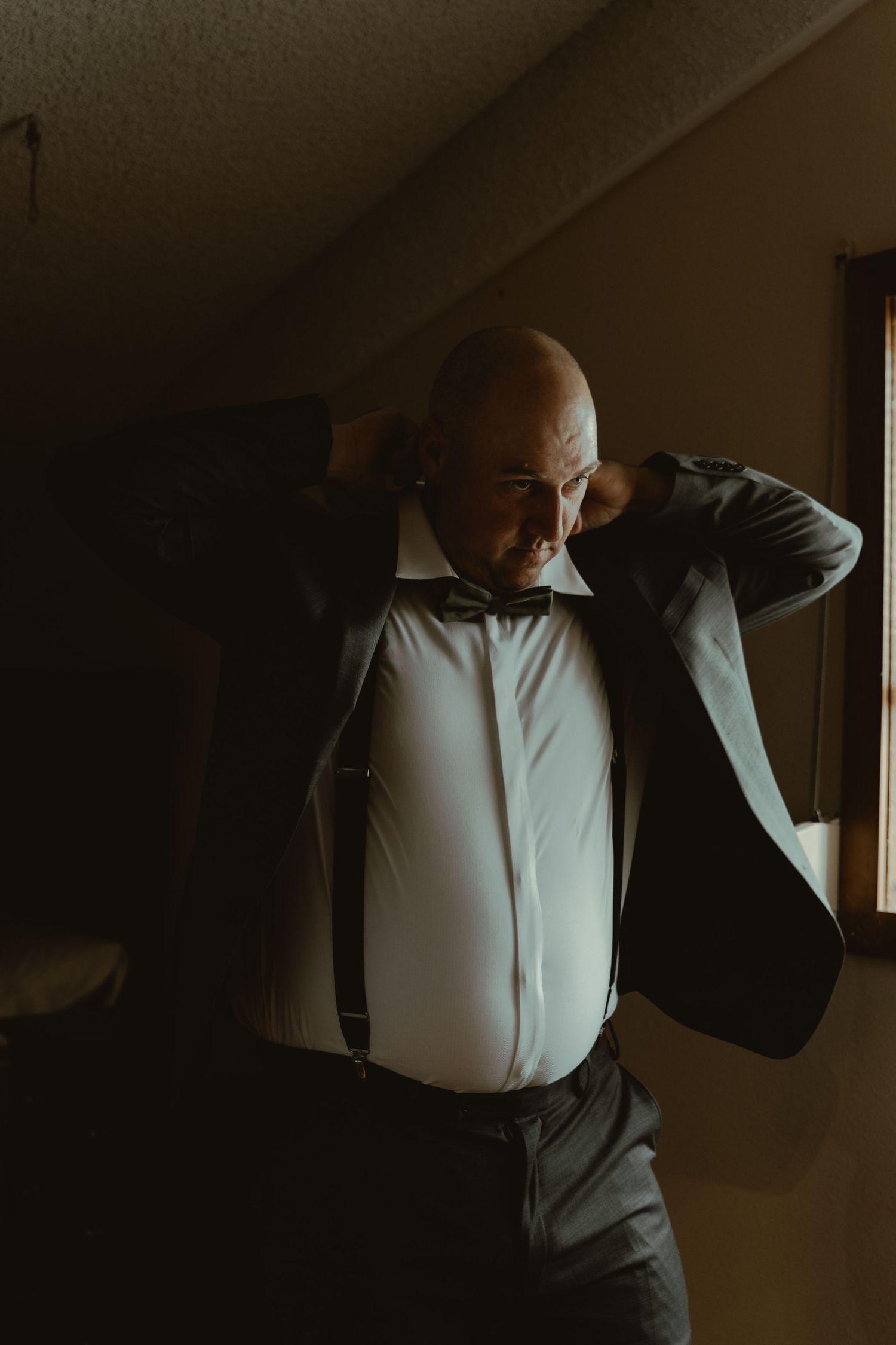 A bald groom wearing suspenders and a bow tie adjusts his dark suit jacket indoors.
