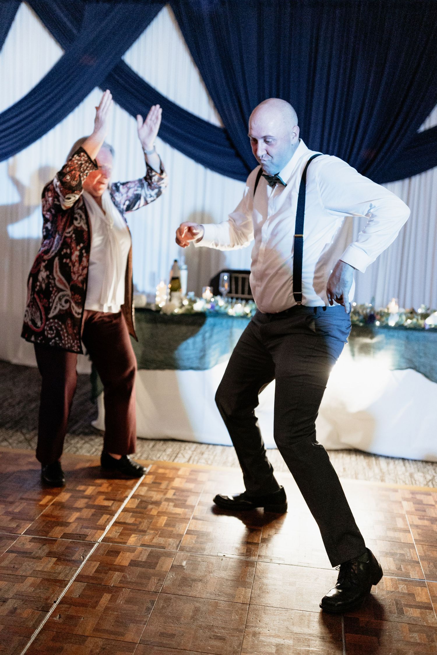 Energetic wedding reception dance floor moment with a bald man in suspenders dancing with an older woman.