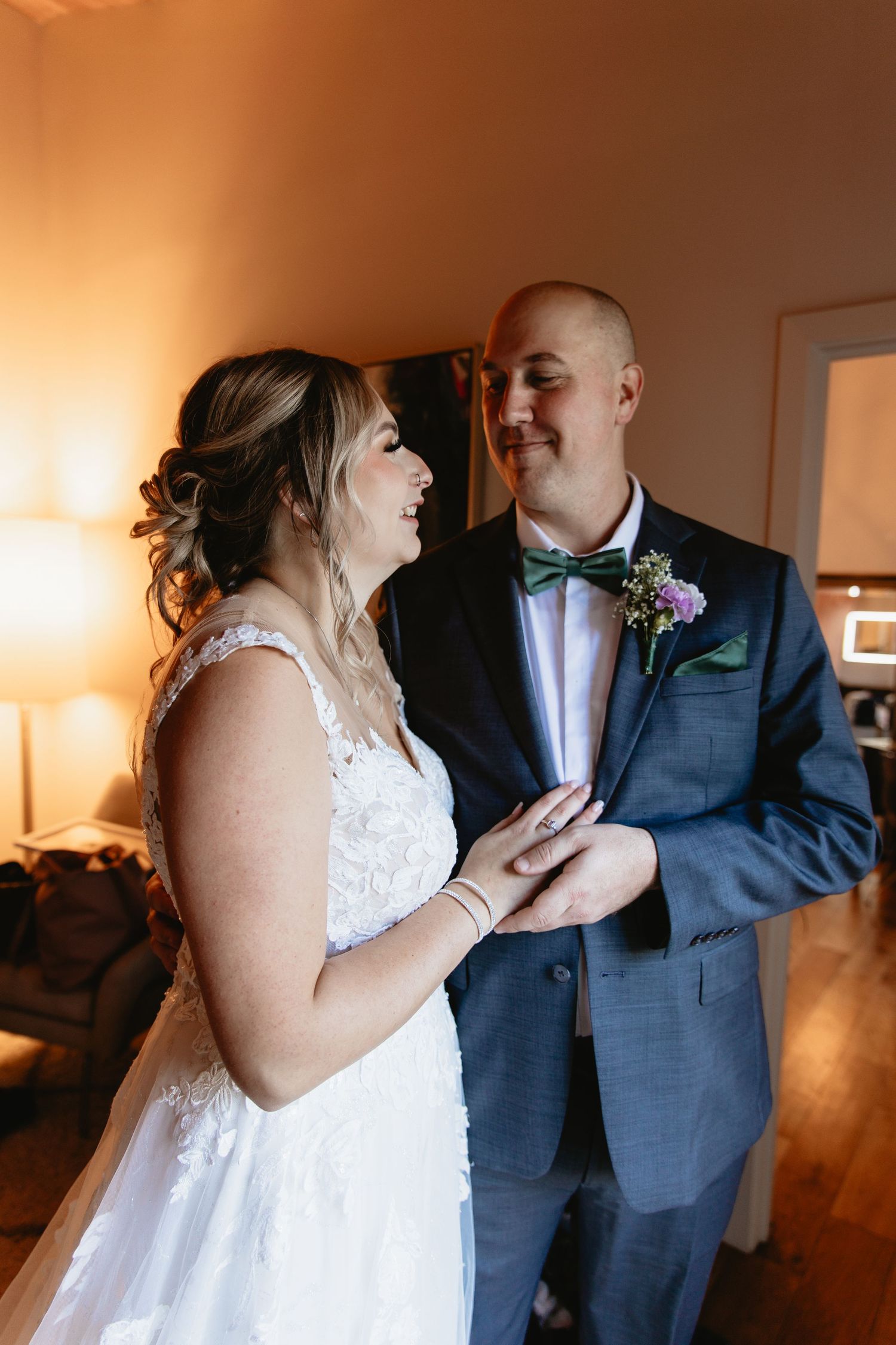 Bride and bald groom in blue suit smiling at each other while holding hands in warm indoor light.