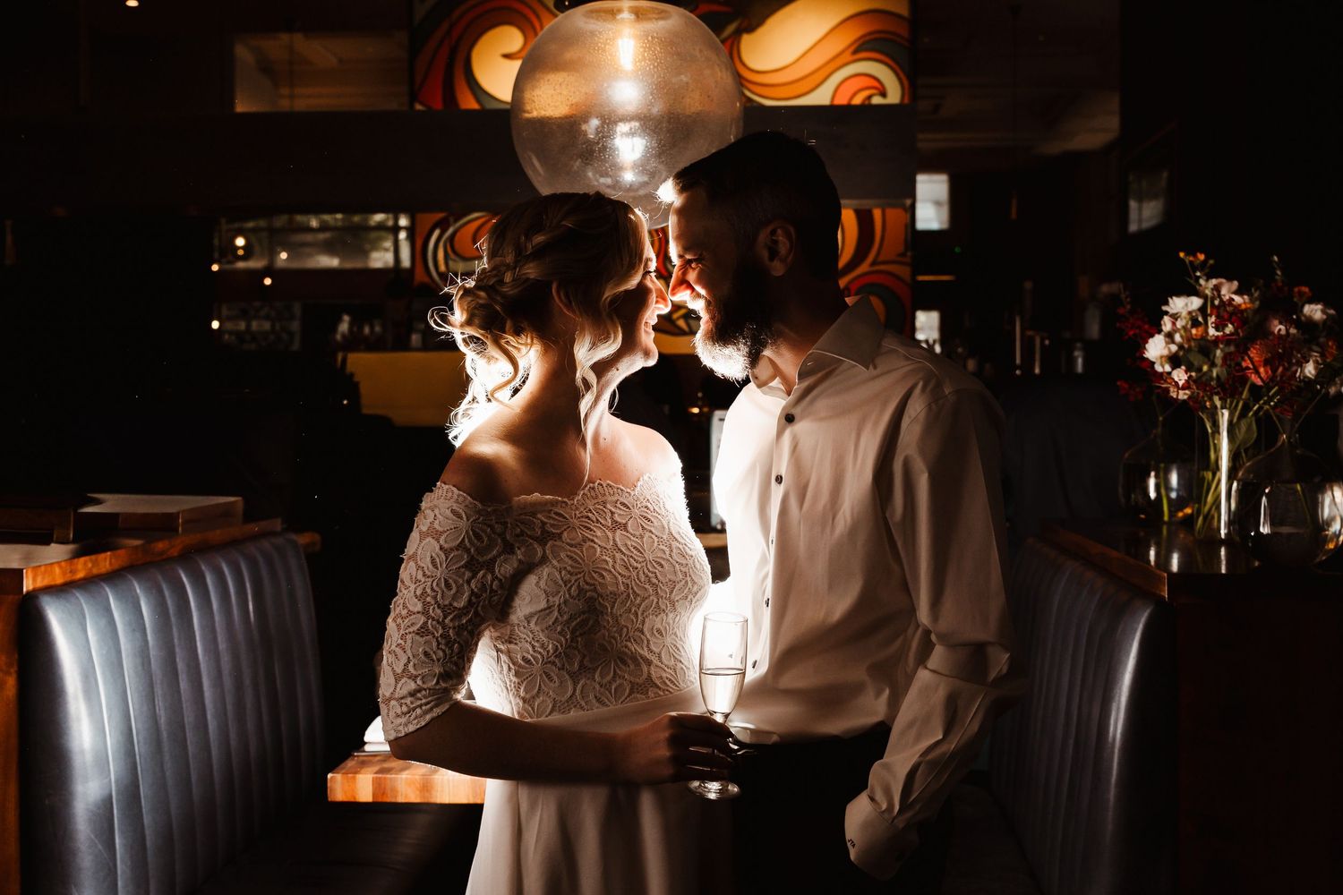 Couple sharing romantic moment at dimly lit bar, bride in lace dress and groom in white shirt, warm golden light.