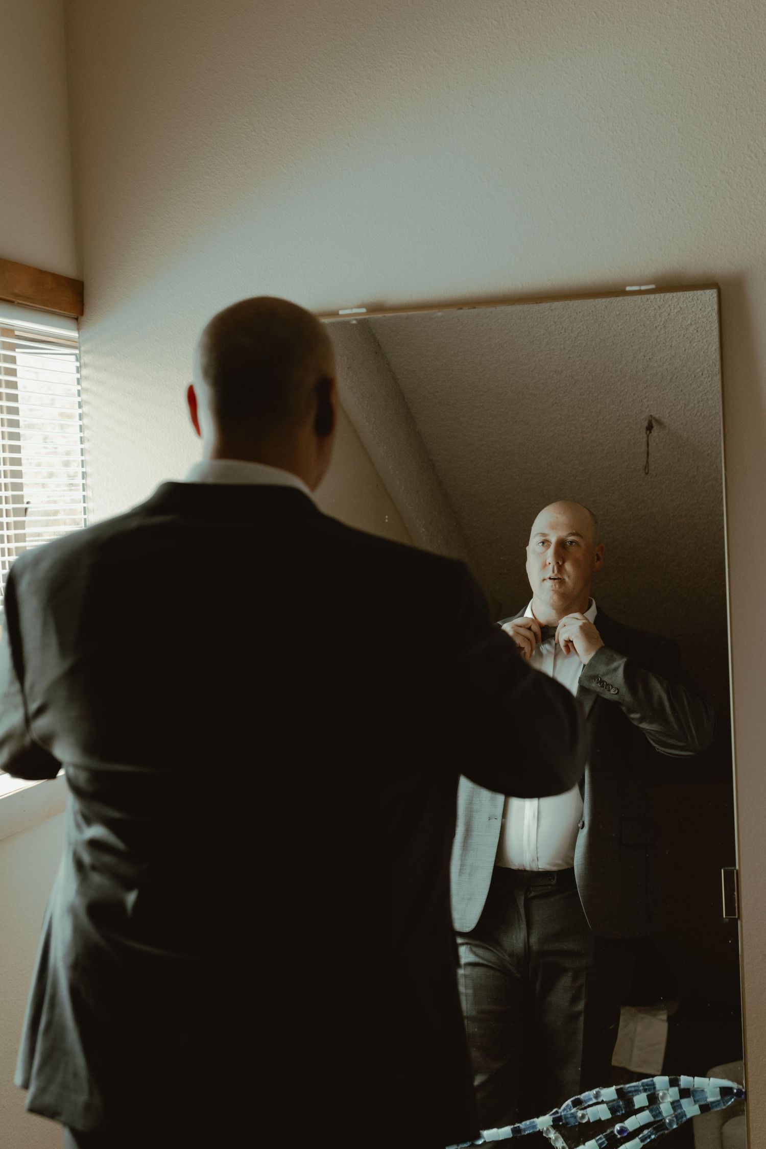 Bald groom in dark suit adjusts his collar while looking at his reflection in a mirror.