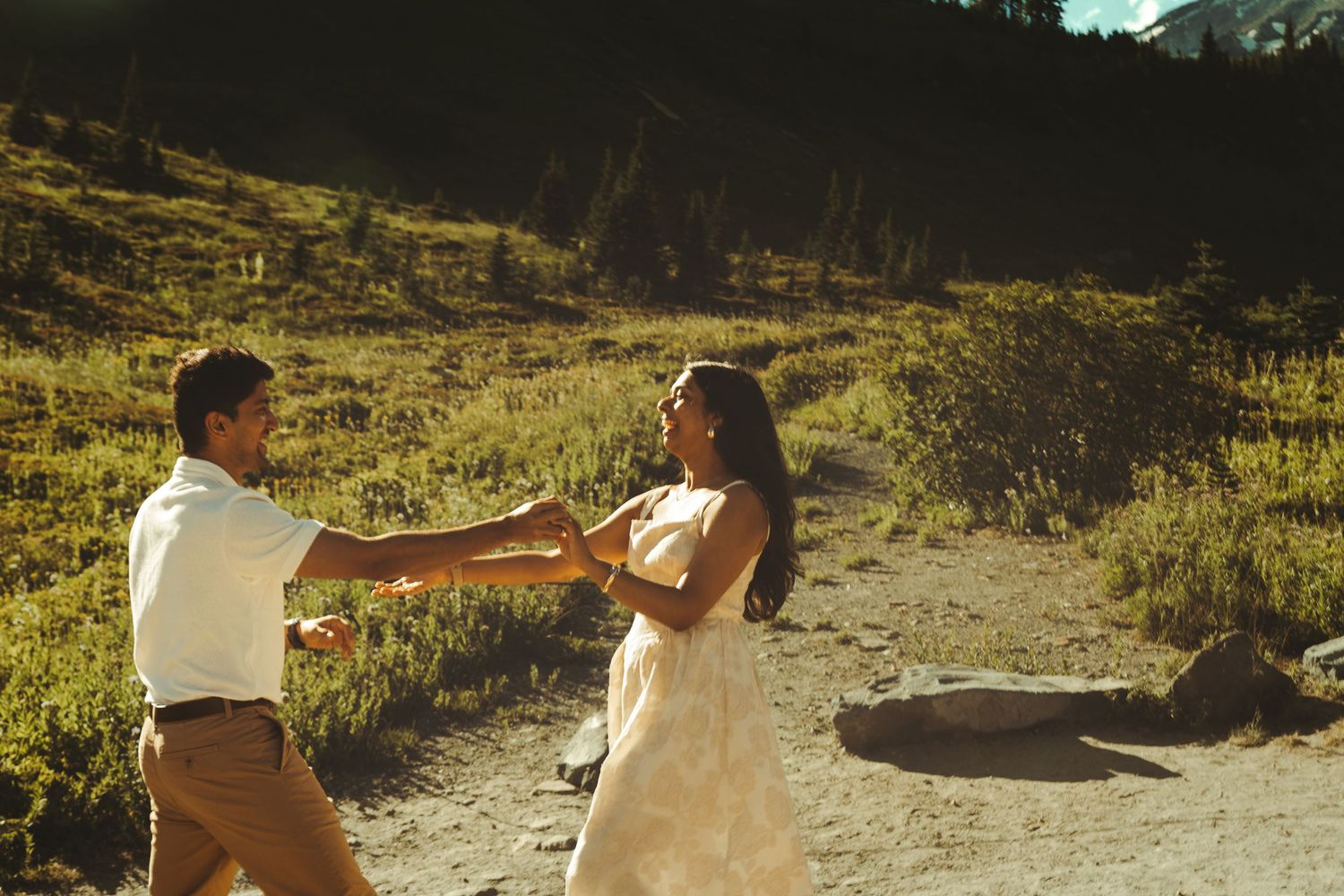 Couple in wedding attire dancing outdoors in a sunlit mountain meadow, holding hands on a dirt path.