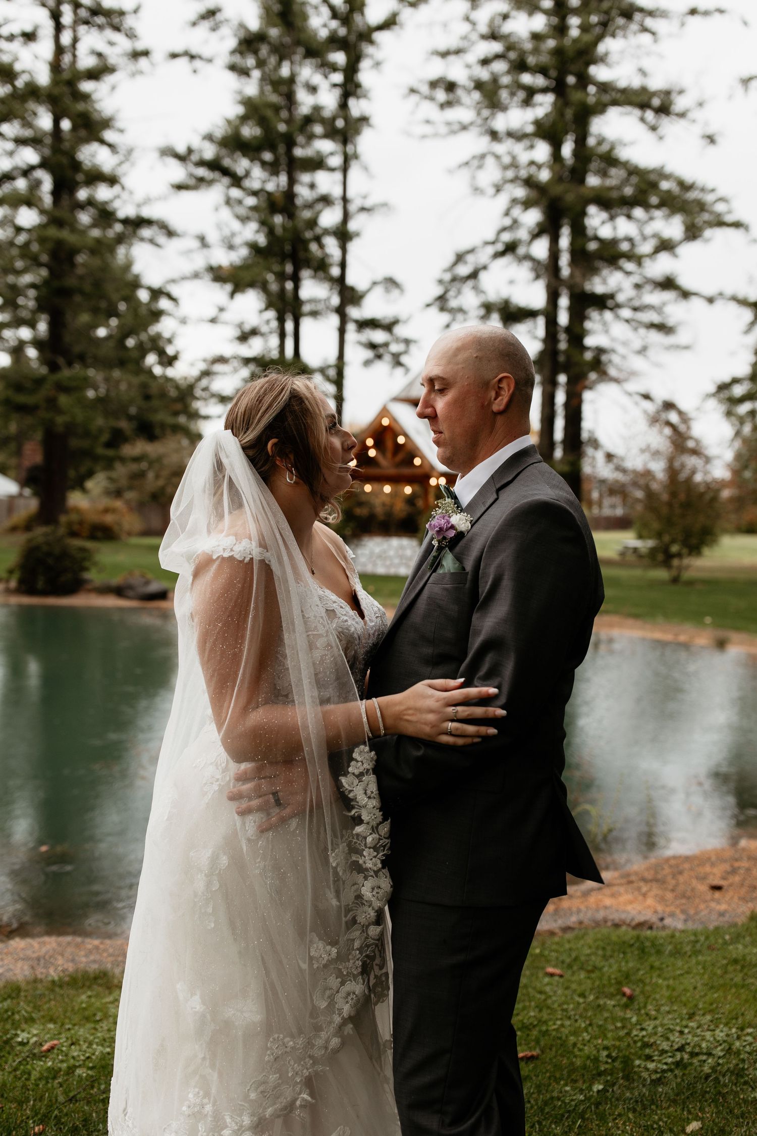 Bride and groom embrace near a pond with tall pine trees backdrop.