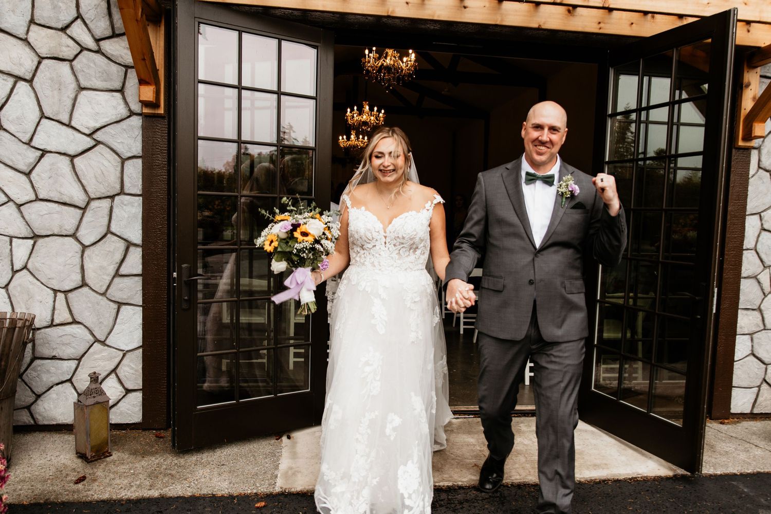 Newlywed couple exits venue through French doors, bride in lace gown holding colorful floral bouquet.