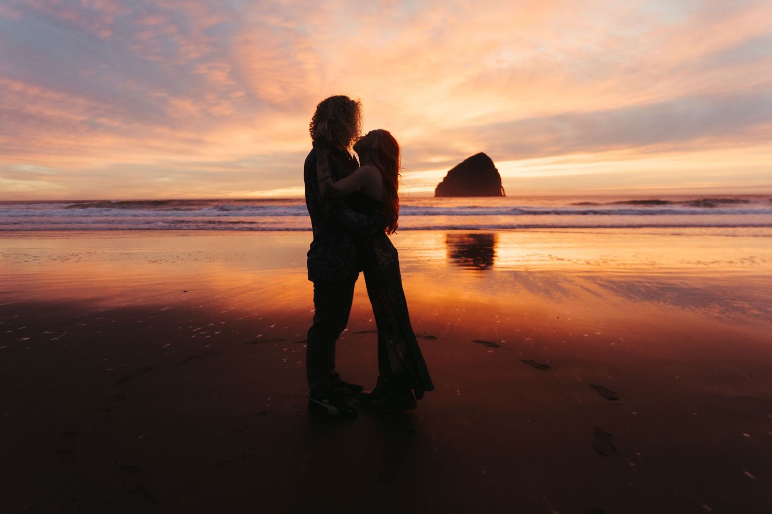 Romantic couple silhouetted against a vibrant pink and orange sunset on a reflective beach.