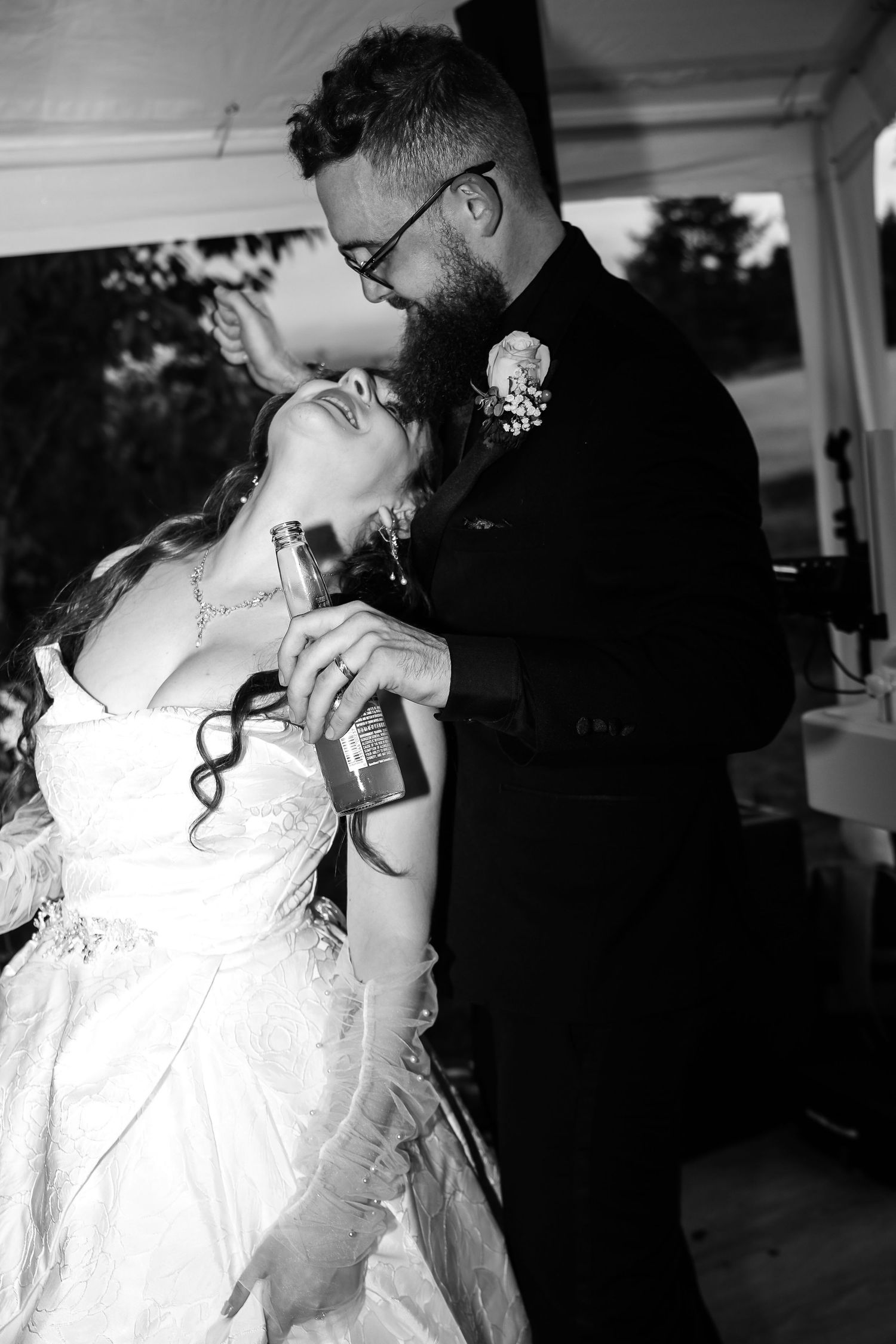 Black and white photo of a bride in a lace gown leaning back as the bearded groom in glasses pours a drink into her mouth at a tent wedding.