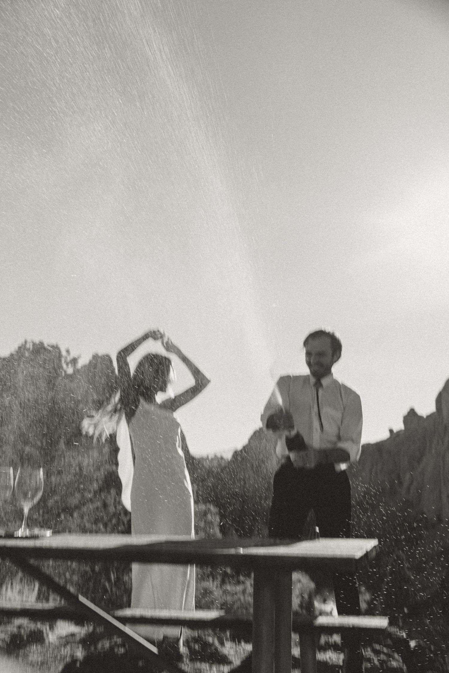 Black and white photo of a couple dancing outdoors near a picnic table, with champagne spraying in the sunlight.