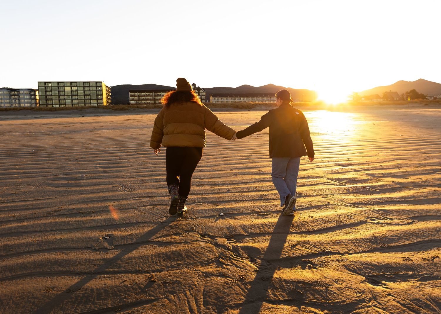Couple holding hands walking on a sandy beach toward a stunning golden sunset.