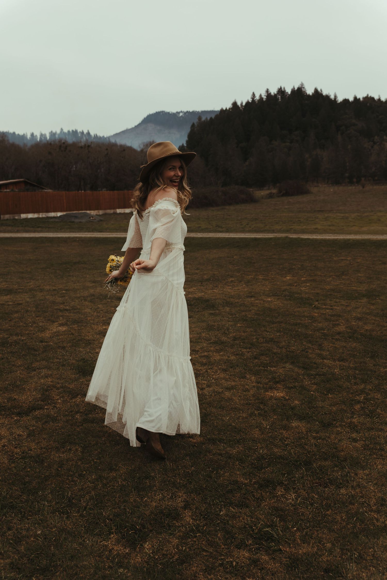 Bohemian bride in white off-shoulder dress and tan hat holding flowers in a scenic mountain meadow.