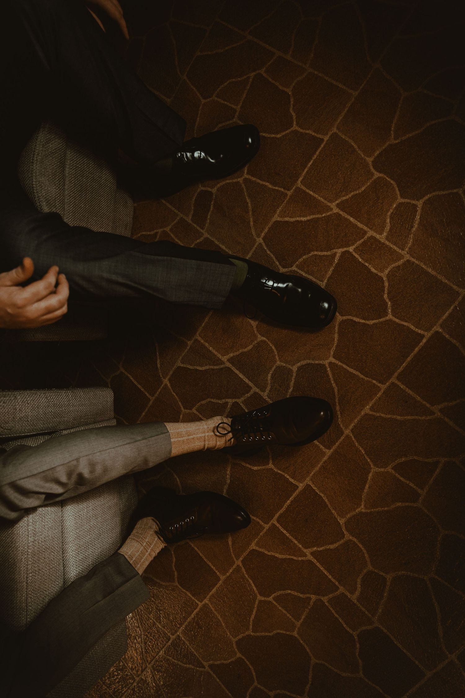 Close-up overhead view of two men in dress shoes and formal trousers seated on chairs.