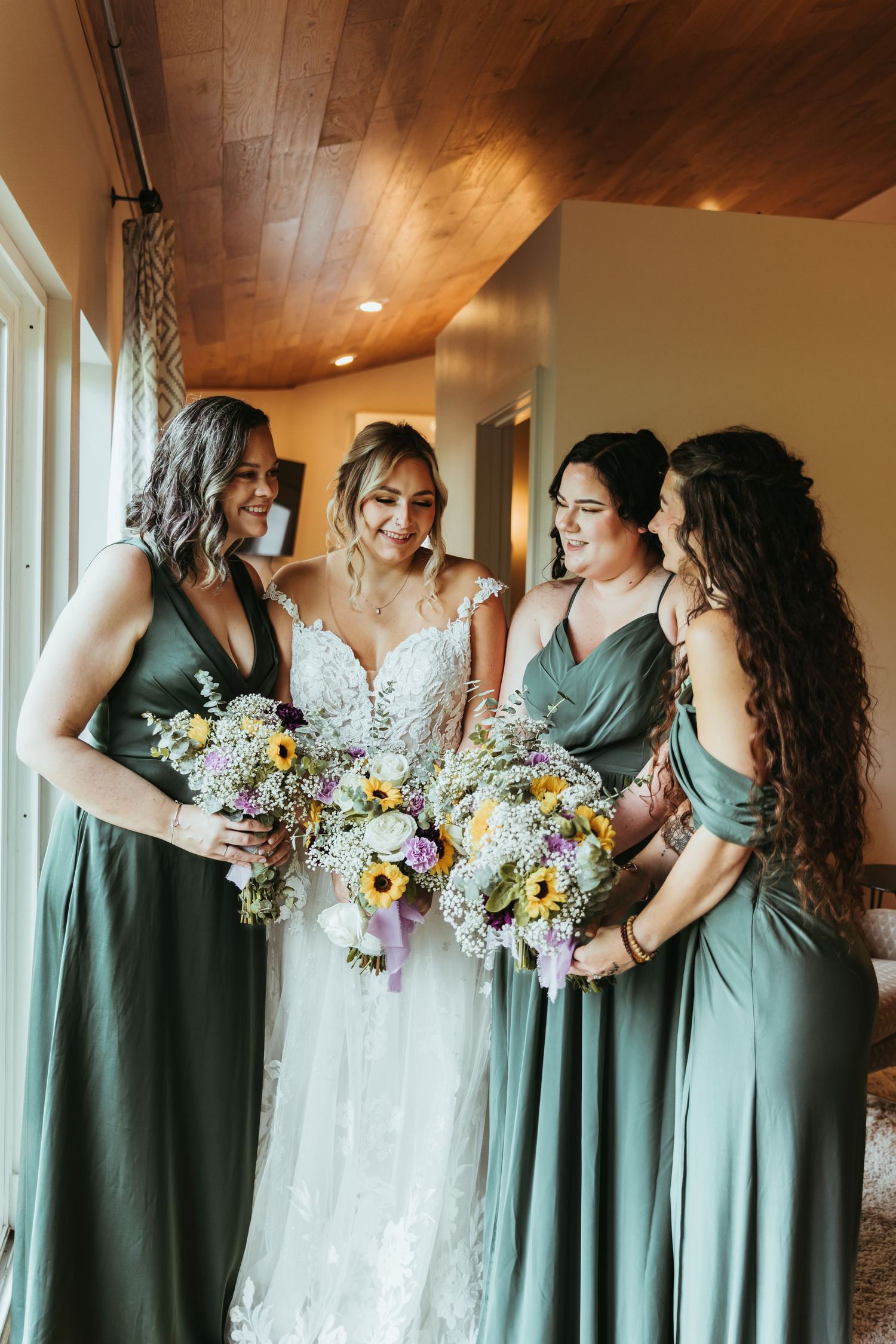 Smiling bride and bridesmaids in matching sage green gowns share a joyful moment holding sunflower bouquets.