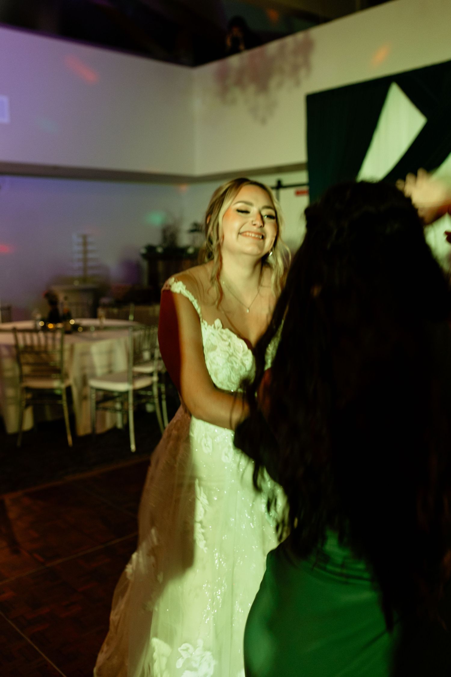 Radiant bride in lace wedding gown smiling and dancing joyfully under colorful lights at the reception.
