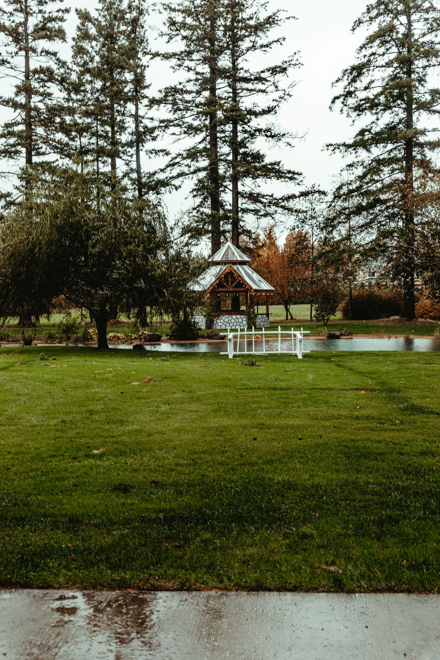 Rainy day outdoor wedding venue featuring a charming gazebo, pond, lush green lawn, and tall pine trees.