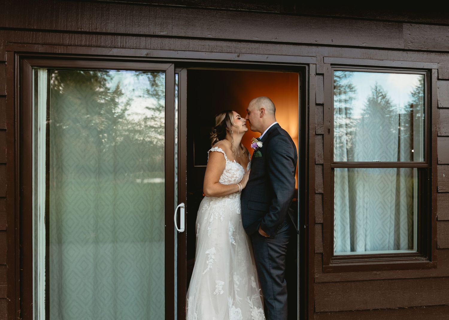 Bride and groom sharing a kiss in the doorway of a brown wooden building on their wedding day.