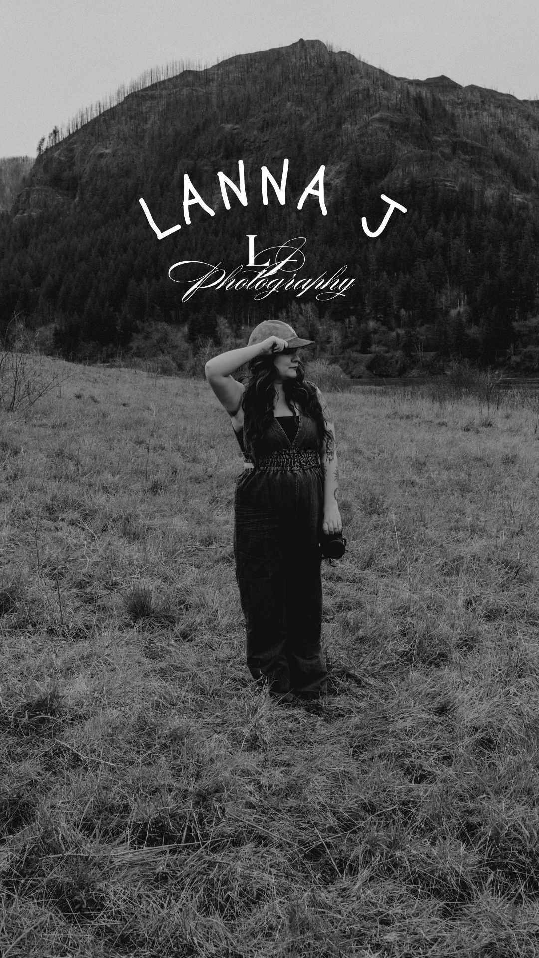 Black and white photo of a woman in a hat and jumpsuit shading her eyes in a grassy field with a mountain, Lanna J Photography logo.