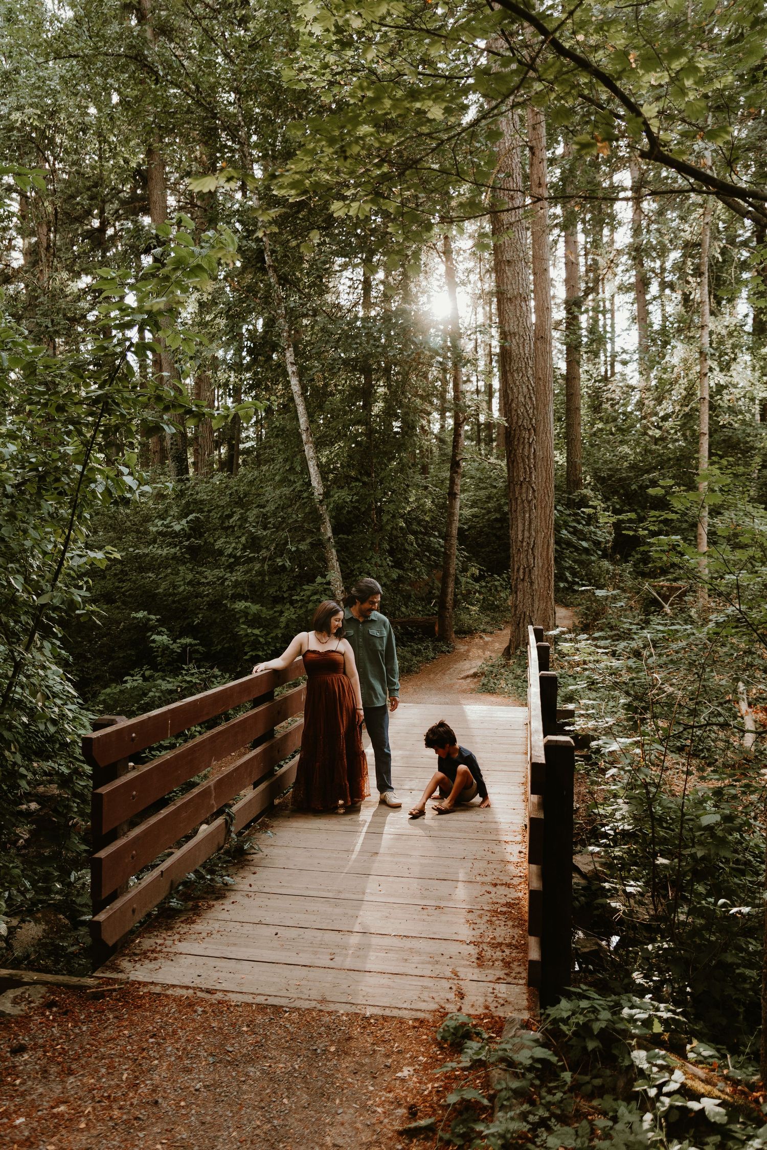 A family stands on a wooden forest bridge surrounded by tall trees, with a child playing on the ground.