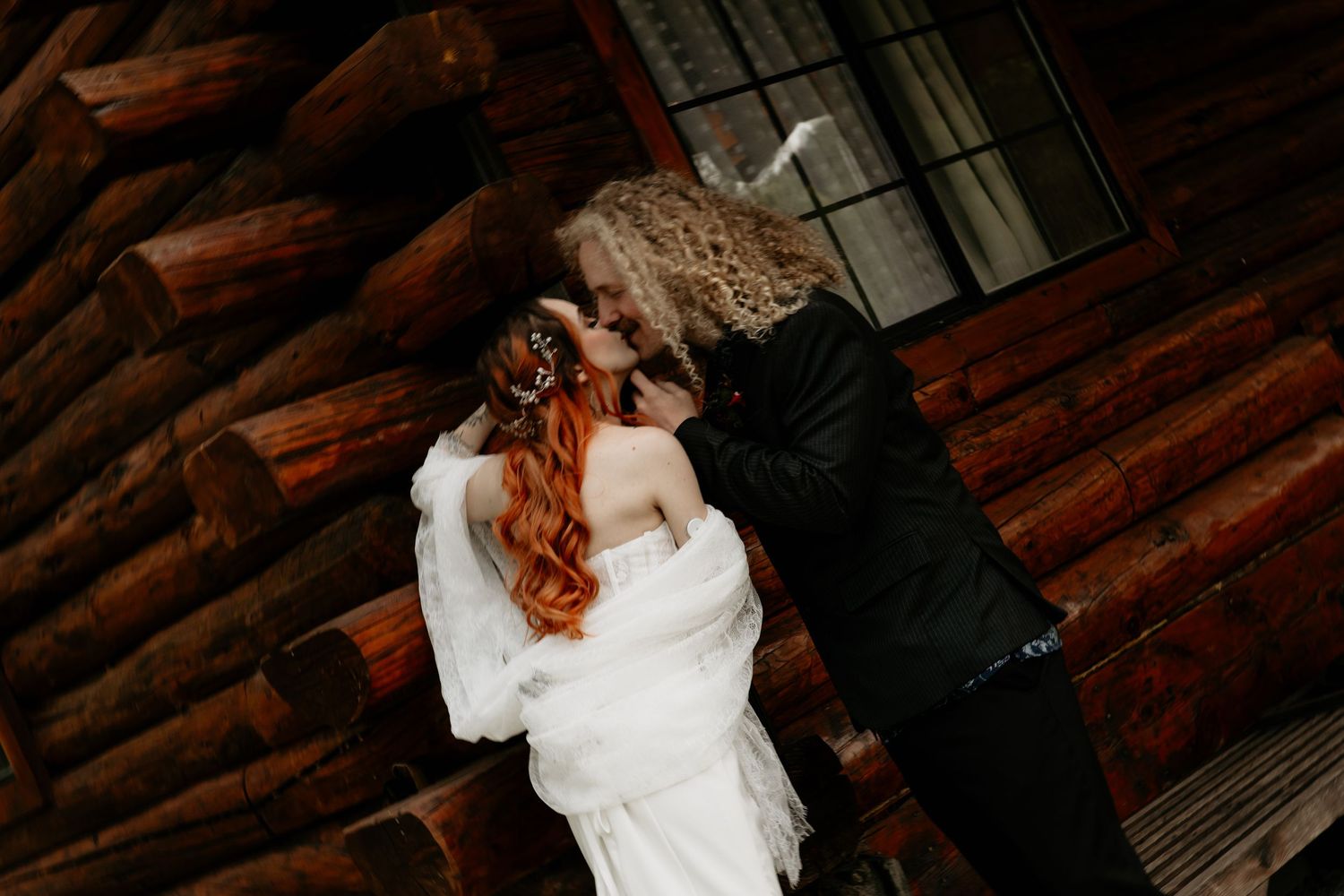 Bride and groom sharing a romantic kiss in front of a rustic log cabin wall on their wedding day.