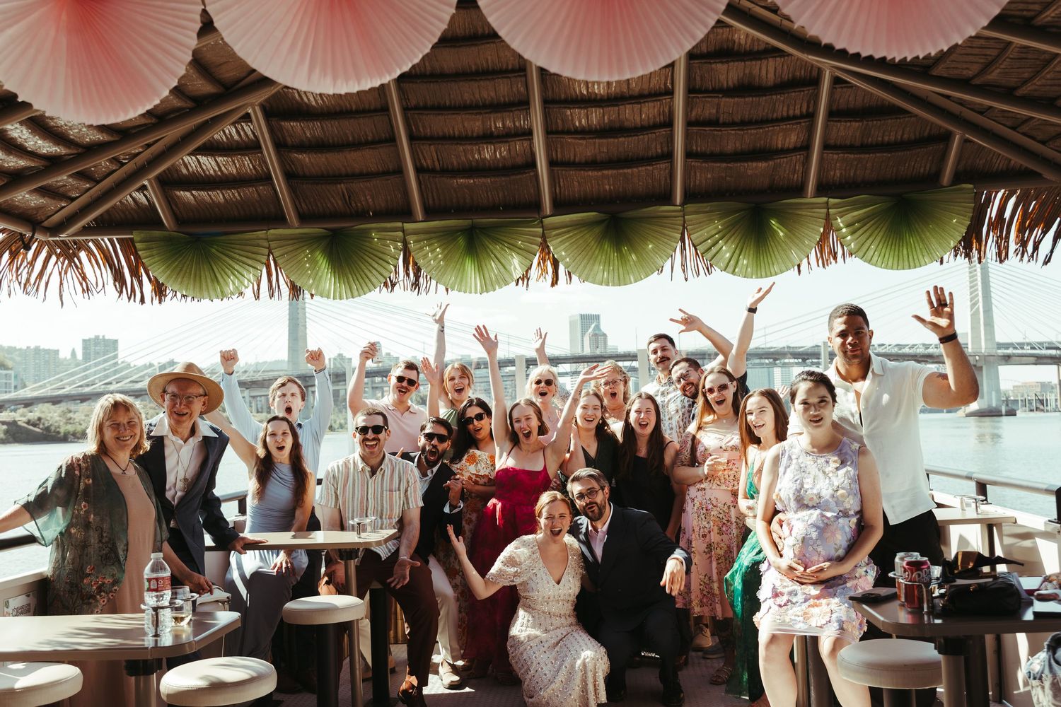 A joyful wedding group photo under a tropical thatched roof with pink paper fans, celebrating by a waterfront venue.