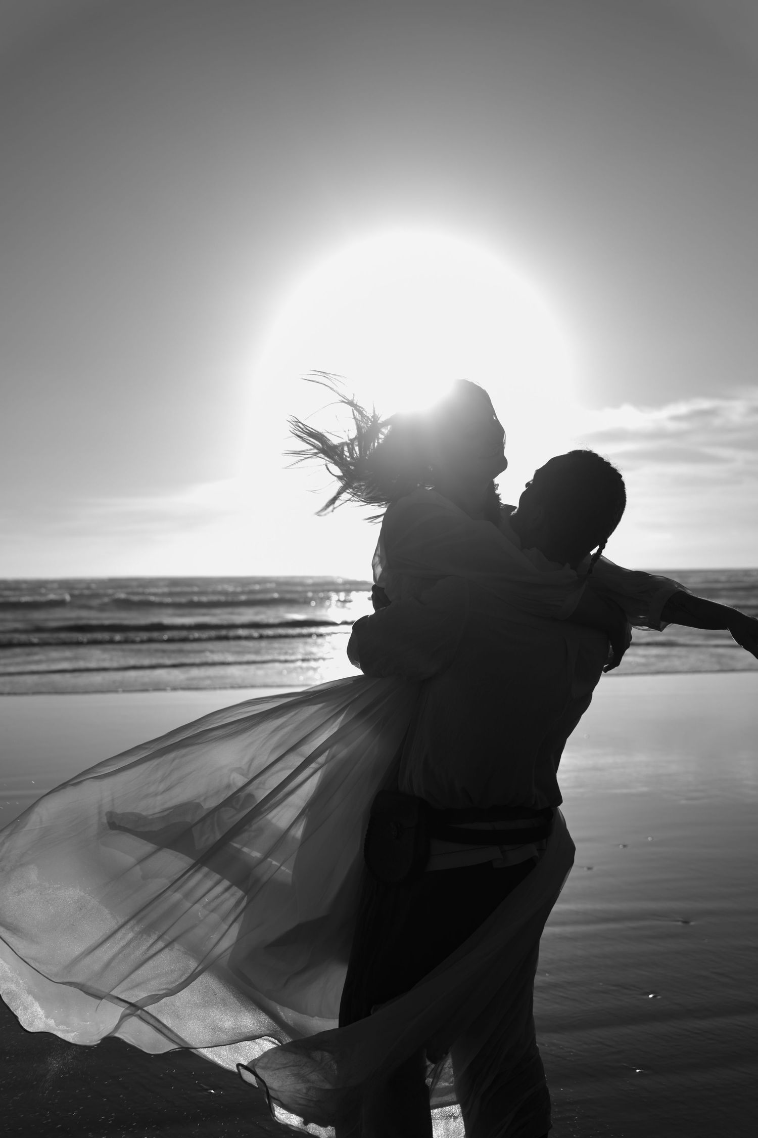 Black and white photo of a couple embracing on a beach at sunset, with a flowing dress billowing in the wind.