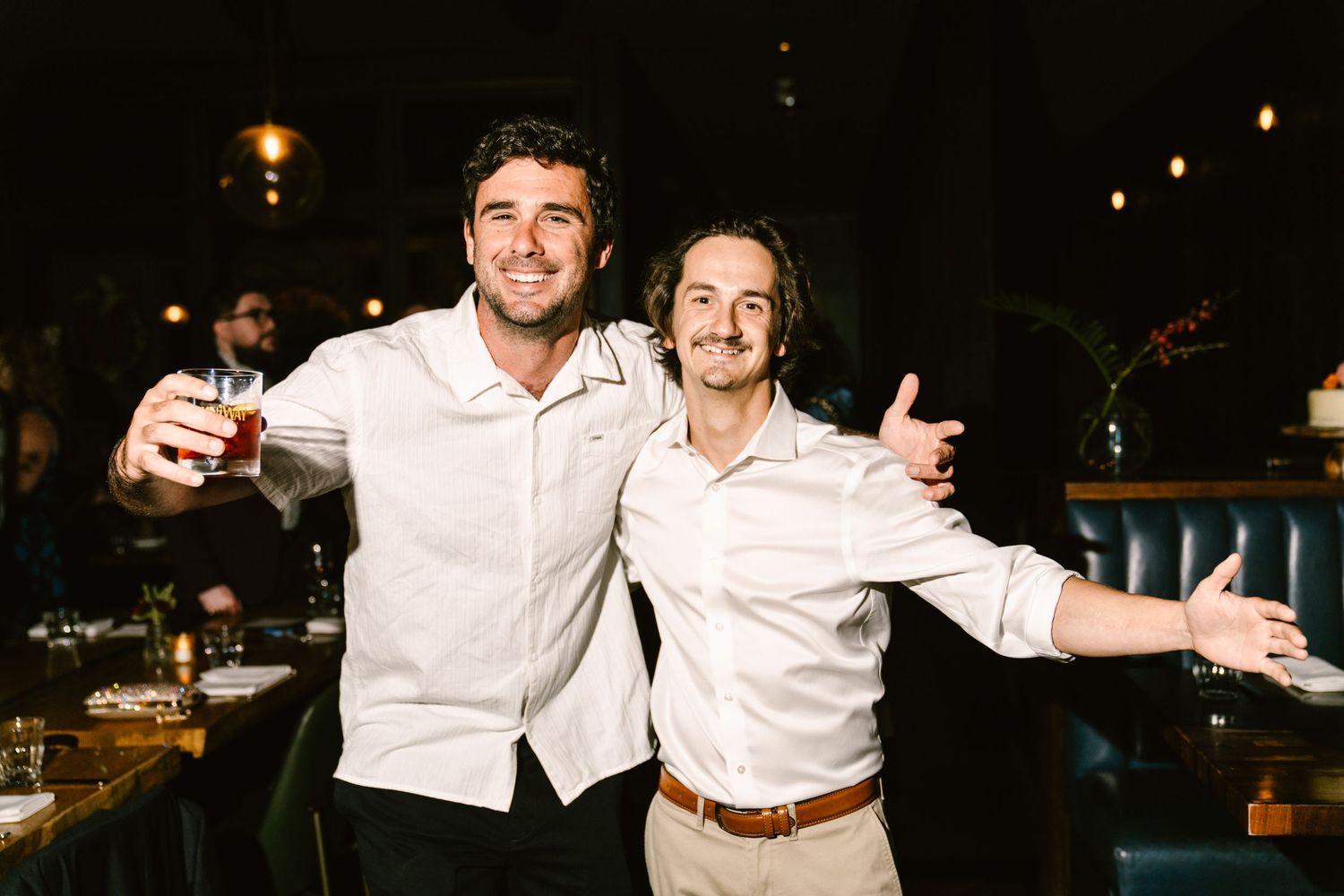 Two smiling men in white shirts celebrate at a bar, one raising a glass and the other spreading arms wide.