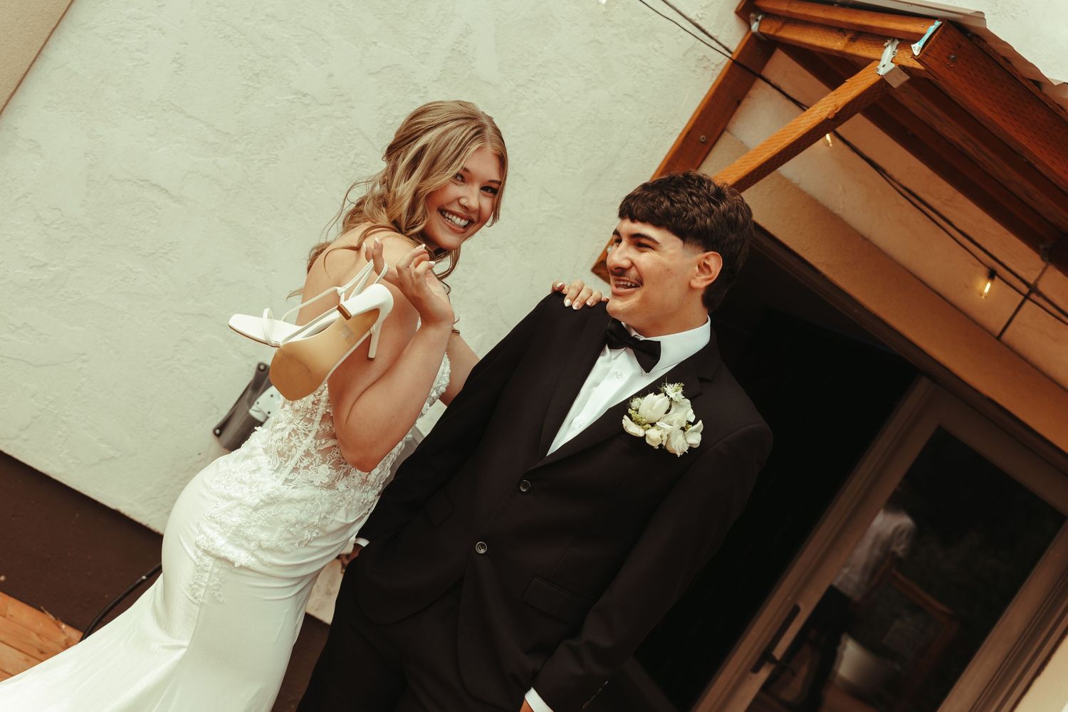Bride in white gown laughing with groom in black tuxedo holding cake knife at wedding reception.
