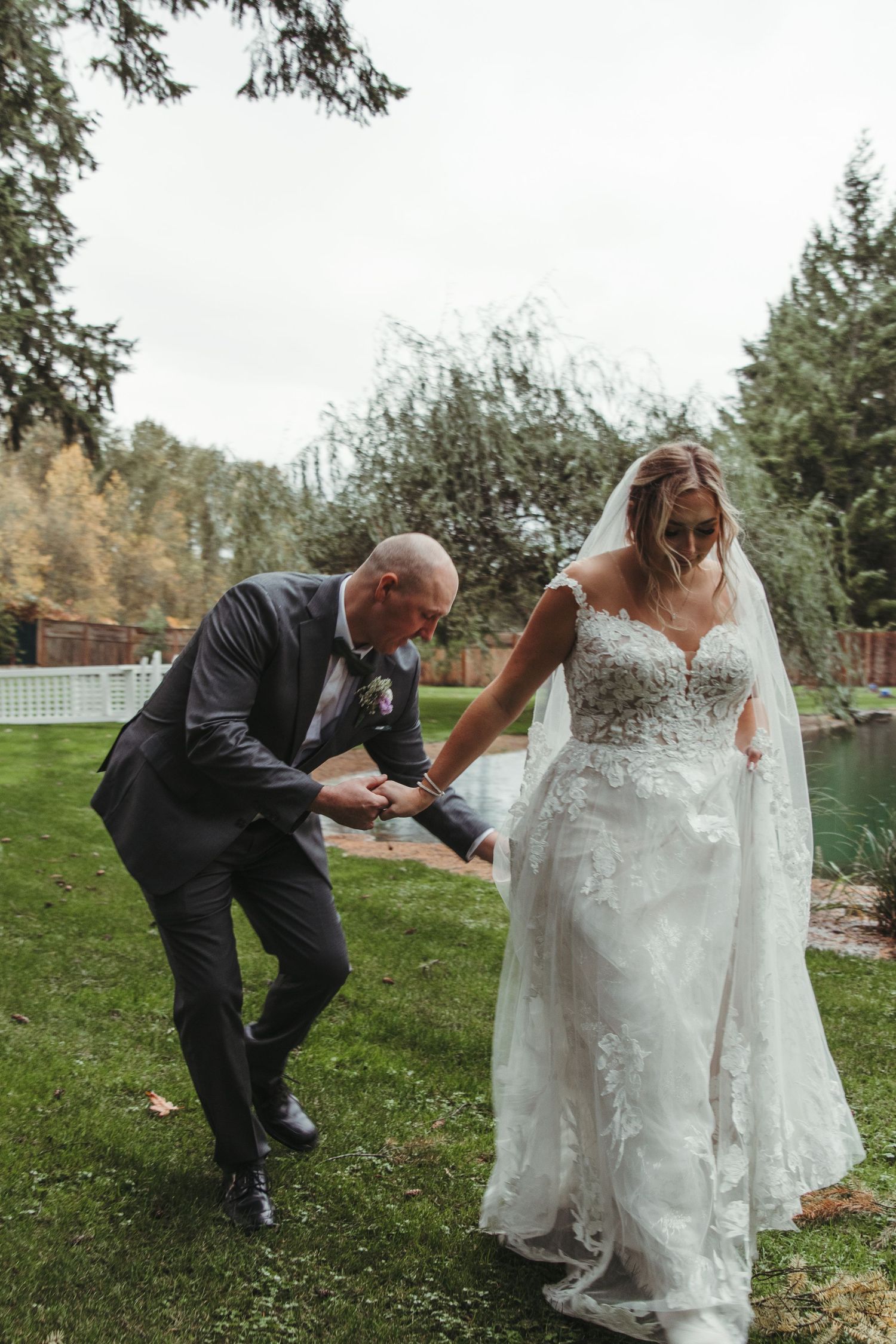 Groom helping bride walk across green grass at an outdoor wedding venue near a pond.