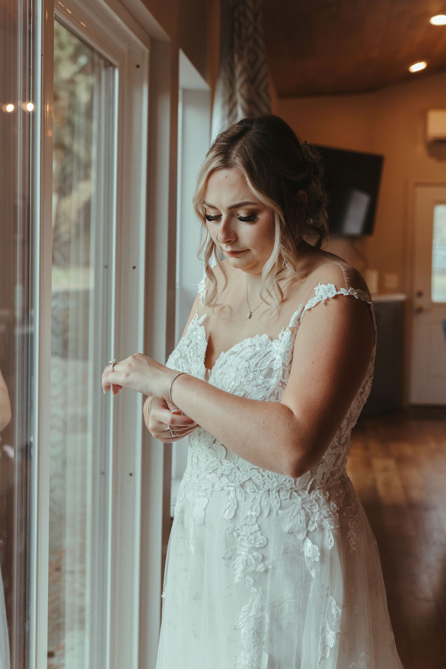 Beautiful bride in lace wedding dress adjusting her bracelet while standing by window.