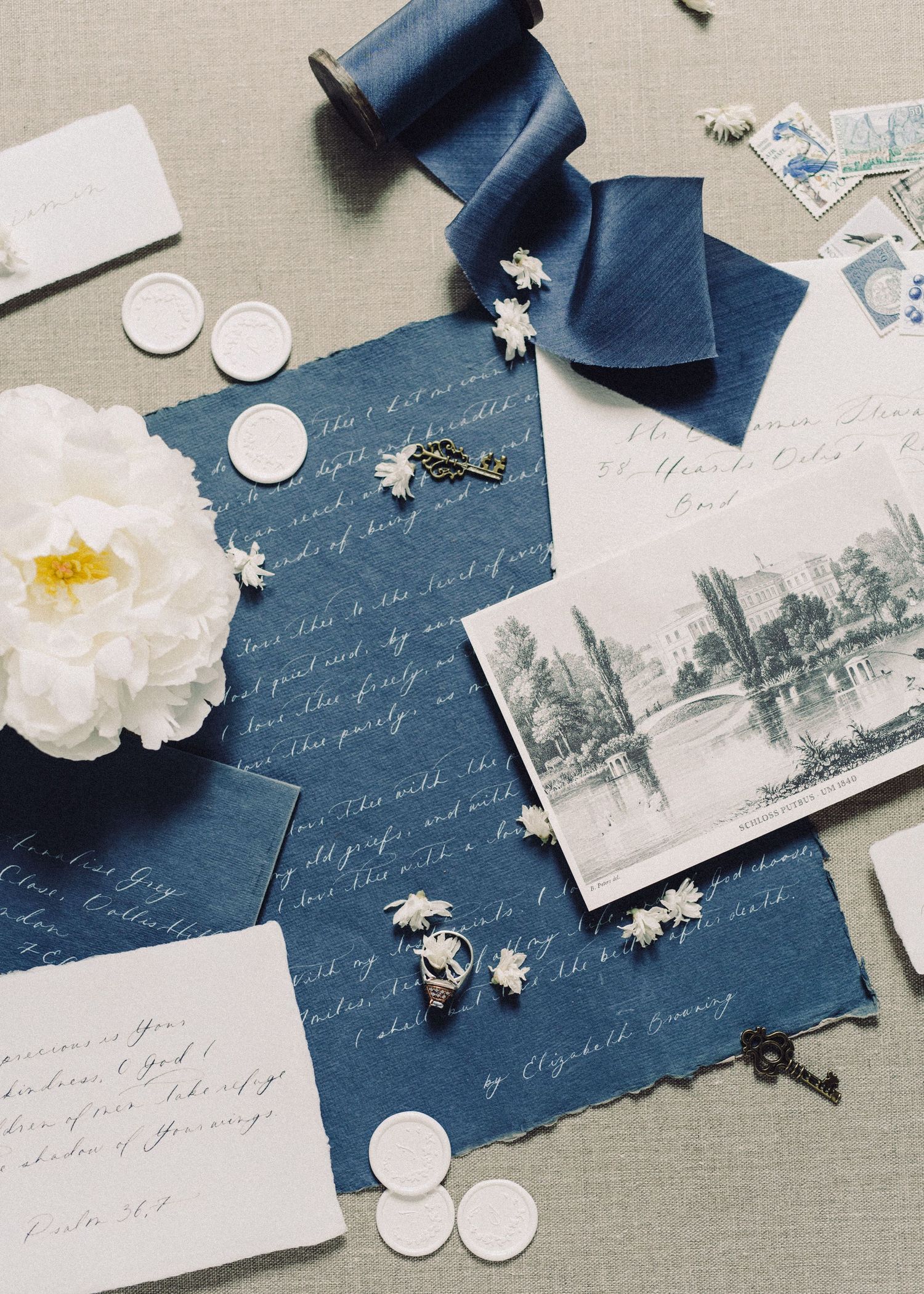 Flat lay of blue wedding stationery with calligraphy, wax seals, silk ribbon, white peony, and vintage postcards.
