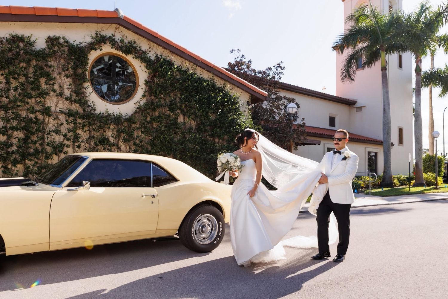 Bride and groom walking past a classic yellow vintage car outside an ivy-covered church with palm trees.
