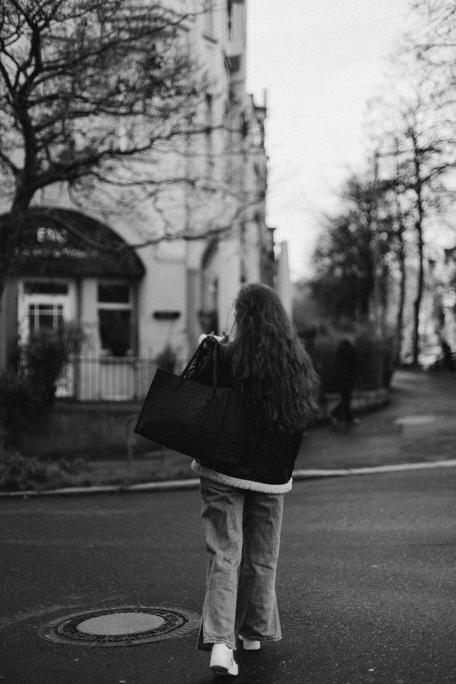 Black and white photo of a woman with long curly hair carrying a large bag on a city street.