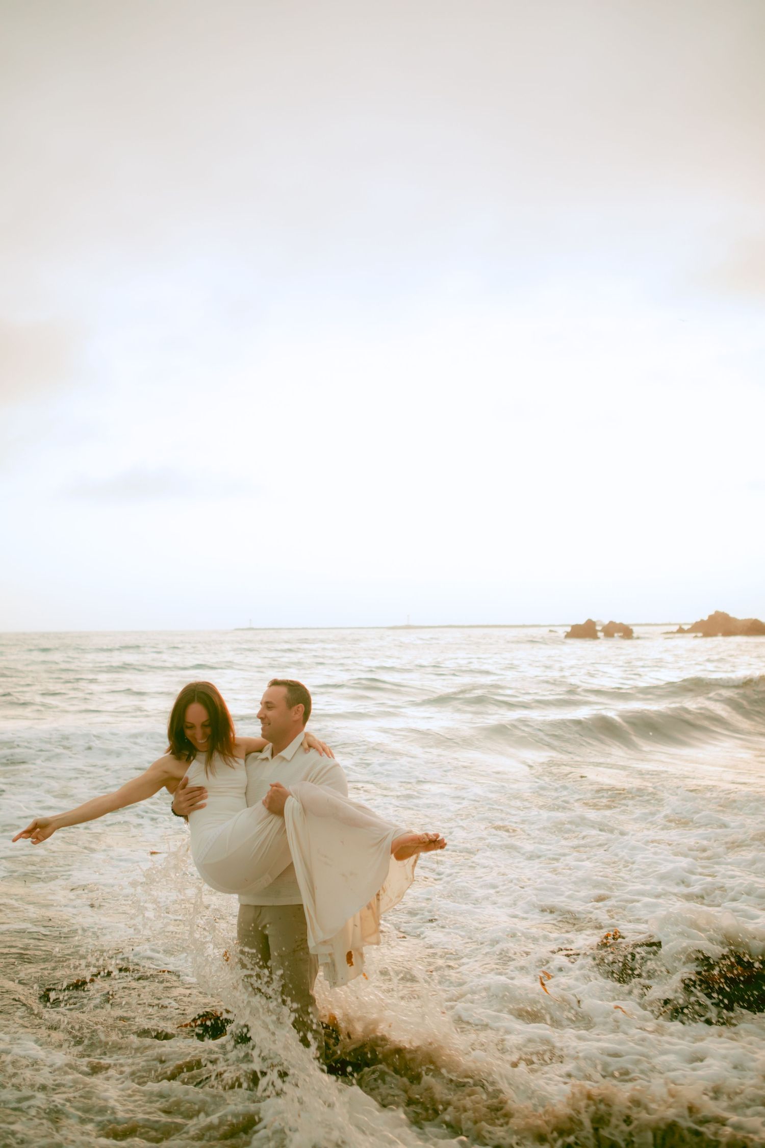 A couple shares a romantic moment on a rocky beach at sunset as ocean waves splash around them.