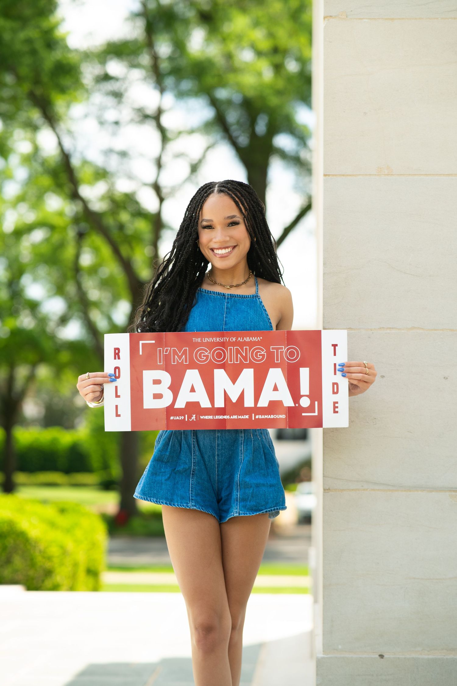 A student in a blue romper holds up a sign celebrating their acceptance to Bama university in an outdoor setting.