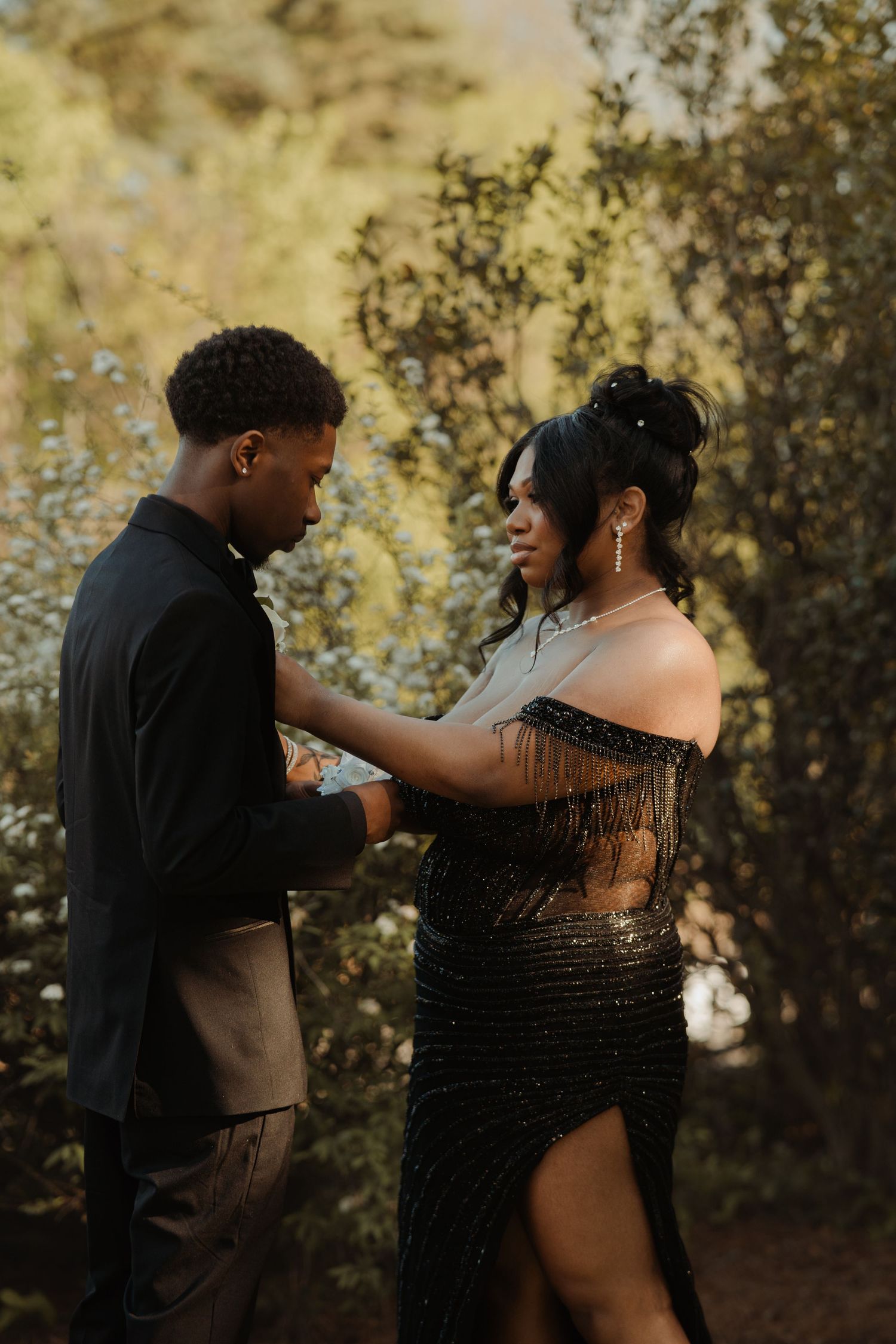 A couple in elegant black attire share an intimate moment during an outdoor photoshoot with golden sunlight.
