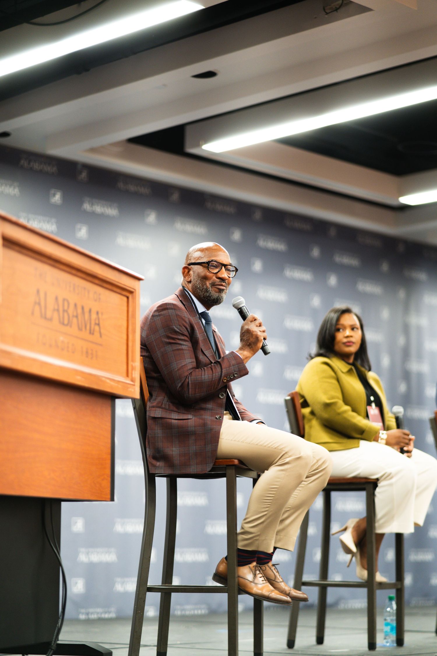 Two speakers sit on tall stools during a professional panel discussion in a conference room setting.